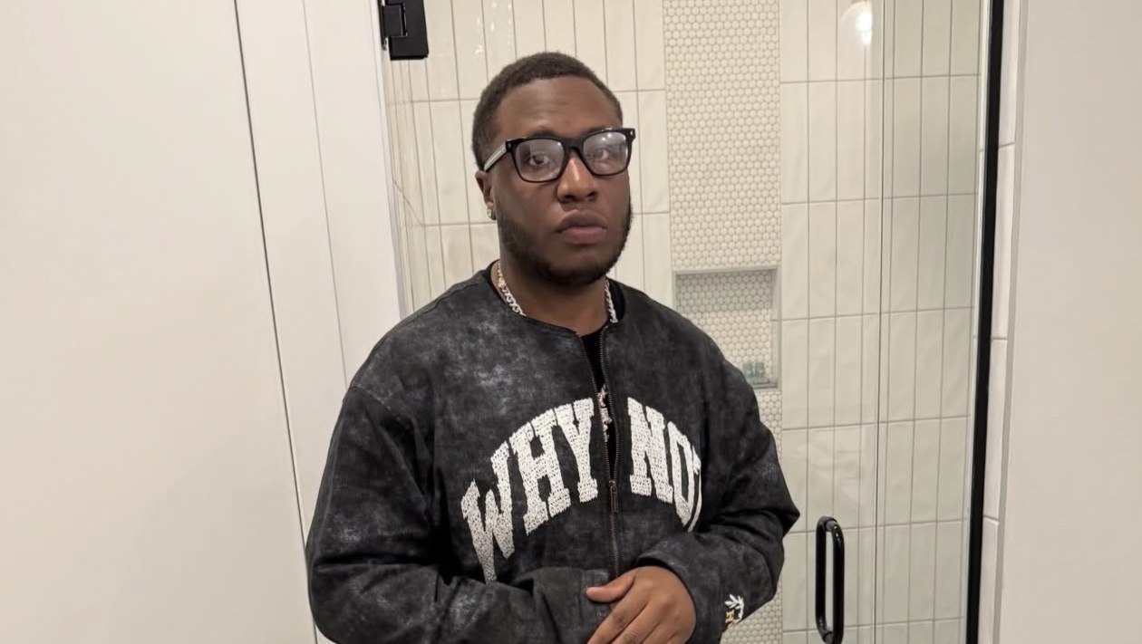 Portrait of Grammy-nominated producer Tay Keith wearing glasses and a dark “WHY NOT” sweatshirt, taken in a modern bathroom setting.